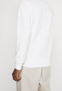 Weißer Sweatshirt mit rundem Halsausschnitt und gerippten Bündchen. Glatte Textur, simples Design, keine sichtbaren Muster. Getragen mit beigen Hosen.