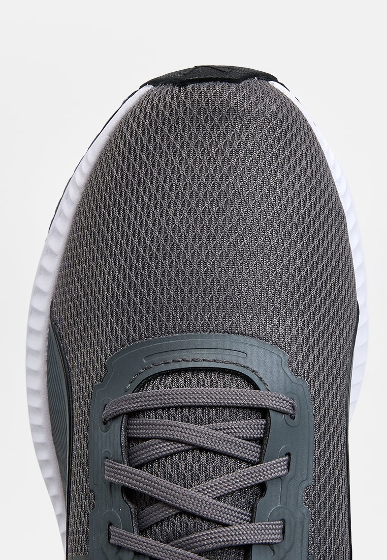 Zapatilla deportiva gris con parte superior de malla texturizada, superposiciones de plástico y una suela blanca acolchada. Cuenta con cordones grises y un diseño de punta redonda.