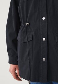 Giacca blu navy con coulisse, hardware argentato, due tasche frontali e bottoni a pressione. Il tessuto sembra resistente con una finitura liscia.