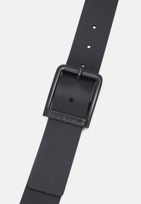 Curea din piele neagră cu o cataramă din metal negru mat. Prezintă găuri pentru ajustare și branding embosat pe cataramă. Textură netedă.
