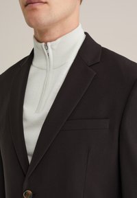 Črn blejzer z strukturirano reverjem, pod katerim se skriva beli pulover s smiselnim zadrgo. Gladko blago, detajl z enim gumbom in žep na prsih.
