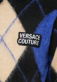 Gebreid sweater met een zwart, blauw en beige ruitpatroon. Inclusief een zwart label met witte tekst: "VERSACE JEANS COUTURE." Zachte textuur.
