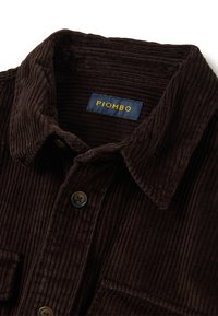 Bruine corduroy shirt met een gestructureerde afwerking, met een klassieke kraag, twee knopen en een marineblauw etiket met de tekst "PIOMBO" in geel.