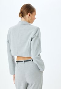 Blazer gris corto con mangas largas, tejido texturizado y solapa con muesca, combinado con pantalones a medida a juego y un cinturón delgado negro.