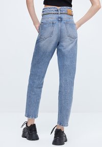 Jeans de cintura alta de color azul claro con una textura desgastada, diseño de cinco bolsillos y un acento de cadena en la cintura. Combinados con zapatillas negras.