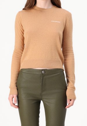 Frau trägt einen beigefarbenen Langarmpullover mit kleinem Text auf der Brust und hoch taillierte olivgrüne Hose vor weißem Hintergrund.