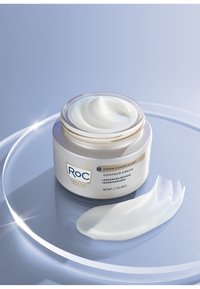 Bocal rond en verre de crème contour blanche avec un léger motif en spirale, étiquettes dorées et bleues. Crème sur une surface circulaire transparente. Capacité de 1,7 oz.