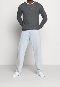 Homme portant un pull texturé gris foncé, un jean bleu clair, des baskets blanches et un collier en chaîne argentée, debout devant un fond clair uni.