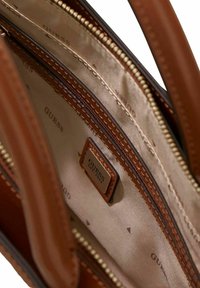 Intérieur d'un sac à main en cuir marron avec une doublure lisse, plusieurs compartiments et une étiquette de la marque GUESS. Détails de fermeture éclair et de couture visibles.