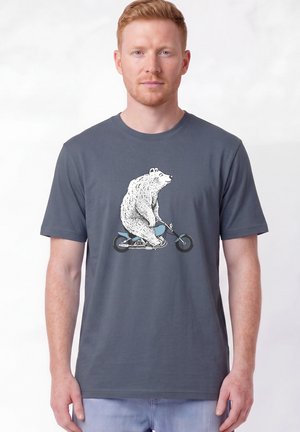 Homme aux cheveux roux courts portant un t-shirt gris avec une illustration d’un ours blanc chevauchant une moto bleue.