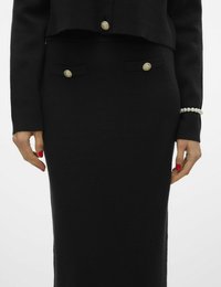 Outfit nero, aderente, che include un giubbotto corto con bottoni dorati e una gonna a tubino elegante con tasche frontali, realizzata in tessuto testurizzato.
