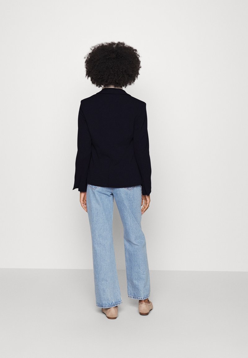 Blazer nero sartoriale con maniche lunghe, abbinato a jeans a gamba larga azzurri. L'outfit presenta un design pulito e minimale senza ulteriori accenti.