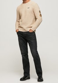 Beige Strickpullover mit strukturiertem Muster, Rundhalsausschnitt und Logo; kombiniert mit dunklen Jeans und schwarzen Stiefeln für einen lässigen Look.