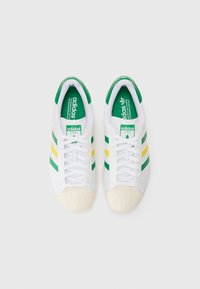White adidas sneakers with green, yellow, and green stripes, rubber shell toe, and green interior. Laced design with a padded collar.