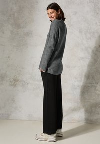 Pull en maille gris à manches longues, avec un ourlet arrondi, associé à un pantalon ample noir et des chaussures de sport beiges. Design minimaliste.