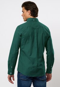 Zöld, hosszú ujjú ing sima anyagból, hegyes gallérral, gombos mandzsettákkal és középen hátsó redővel a nagyobb dizájn részletért.