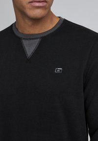 Schwarzer Langarm-Sweatshirt mit geripptem Ausschnitt, kontrastierendem grauen Akzent unter dem Kragen und gesticktem Logo auf der Brust.