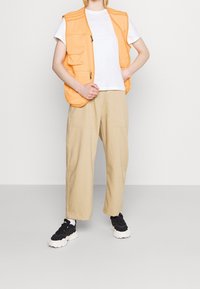 Orange väst med flera fickor över en vit t-shirt, kombinerad med beige vida byxor och svarta sneakers.