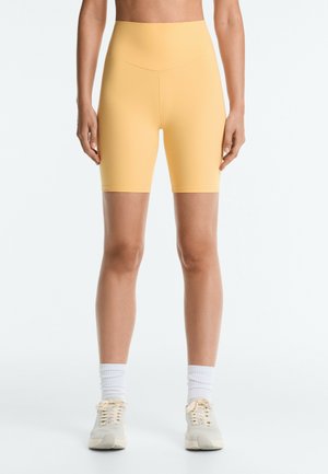 Personne portant un short cycliste jaune taille haute, des chaussettes blanches et des chaussures de sport beiges, debout devant un fond uni clair.