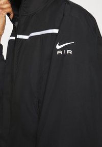 Veste Nike noire avec fermeture éclair, bande réfléchissante blanche traversant la poitrine, et logo imprimé « AIR » sur la manche. Texture de tissu lisse.