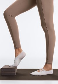 Personne portant des leggings beiges et des chaussettes blanches antidérapantes, étirant le pied sur des blocs de yoga empilés sur un tapis de sport marron.