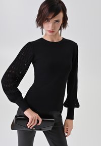 Schwarzer Strickpullover mit eng anliegendem Körper und strukturierten, offen gestrickten puffärmeln. Kombiniert mit einer schwarzen Lack-Clutch, die ein goldenes Logodetail aufweist.