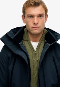 Chaqueta azul marino oscura con un alto cuello y cremalleras, superpuesta a un suéter de punto trenzado verde y una camisa blanca. Diseño limpio y moderno.