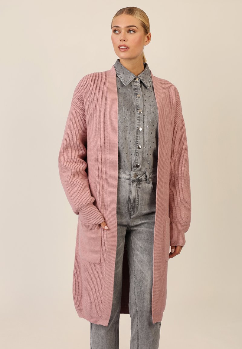 Lang roze cardigan van ribgebreide stof, met een open voorkant, lange mouwen en twee zakken aan de voorkant. Gekozen boven een spijkerblouse.