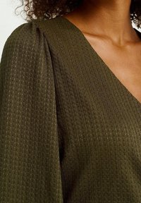 Olivgrön topp i ett strukturerat vävt tyg, med V-ringning och puffärmar. Närbild visar det detaljerade mönstret och glansen.