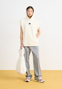 Beige ärmelloser Hoodie mit Kapuze, schwarzen Akzenten, grauen weit geschnittenen Hosen, hält eine strukturierte weiße Tasche und trägt weiße Sneakers mit roten Akzenten.