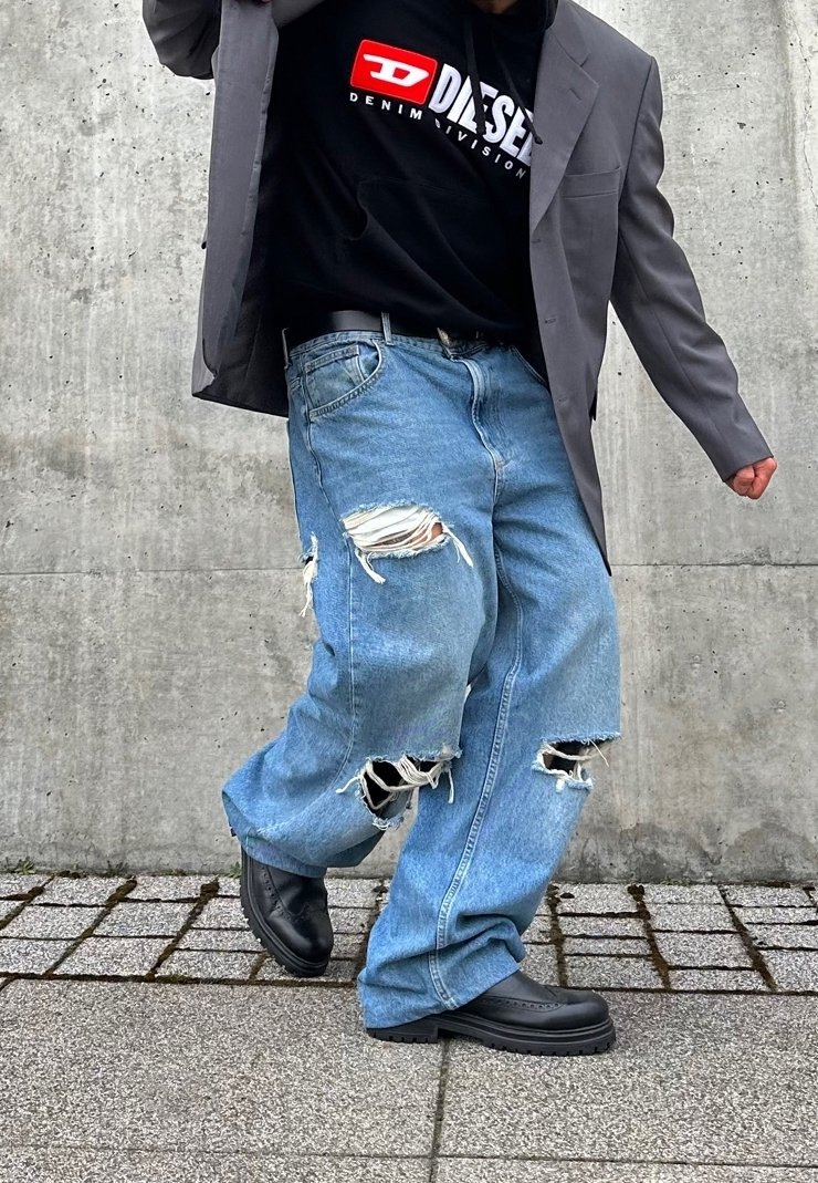 Personne portant un jean large déchiré, un sweat à capuche Diesel noir, un blazer gris et des chaussures noires, debout sur un trottoir pavé contre un mur en béton.