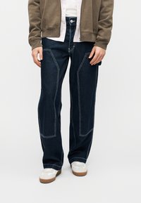 Σκούρα μπλε τζιν με άνετη εφαρμογή και χαρακτηριστικές γωνιώδεις ραφές. Φορεμένα με γκρι φούτερ με φερμουάρ και λευκά αθλητικά παπούτσια.