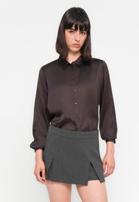 Une jeune femme aux cheveux foncés et raides porte une blouse boutonnée marron foncé et une mini-jupe grise avec des plis à l'avant, se tenant contre un fond blanc.