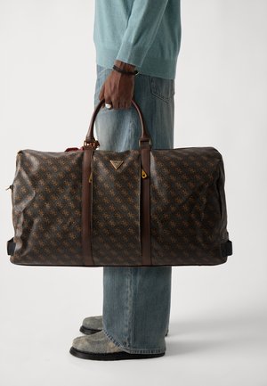 Borsa a duffel marrone con motivo, dotata di manici in pelle scura e dettagli dorati, caratterizzata da un design spazioso, portata da una persona in abbigliamento casual.