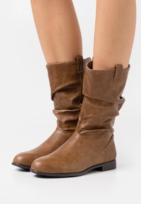 Bottes cheville en similicuir marron à design décontracté, bout arrondi, semelle plate et languettes sur le dessus pour faciliter l'enfilage.