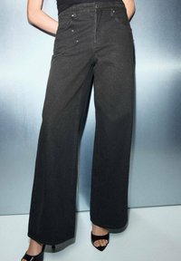 Jeans de pierna ancha negros, confeccionados en denim con un acabado texturizado y sutiles adornos. Presentan bolsillos clásicos y una cinturilla con trabillas para cinturón.
