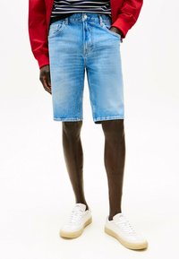 Bermuda in denim azzurro chiaro con un design classico, tasche frontali e orli rifiniti, abbinate a sneakers bianche. Giacca rossa sopra.