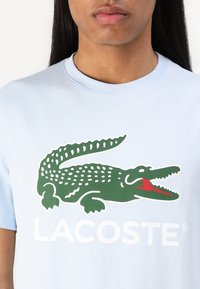 Šviesiai mėlynas medvilnės marškinėlis su dideliu žaliu krokodilo piešiniu, turinčiu raudonus akcentus ir baltu "LACOSTE" tekstu apačioje.