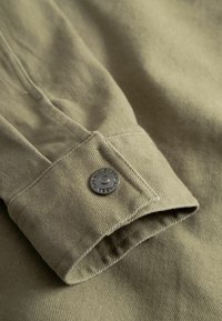 Olivengrønne manchetærmer med lange ærmer og en metaltrykknap graveret med mærkenavnet, sat mod en matchende stofbaggrund.