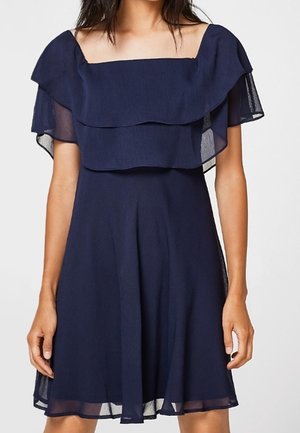 Femme portant une robe bleu marine à épaules dénudées avec des volants superposés et des manches courtes transparentes.