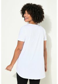 T-shirt blanc à manches courtes avec un ourlet arrondi, fabriqué en tissu doux. Le modèle a des cheveux bouclés et se tourne légèrement vers l'arrière.