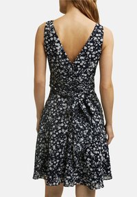 Kvinna som bär en ärmlös marinblå klänning med vit blommig mönster, V-rygg och en knuten rosett vid midjan, sedd bakifrån.