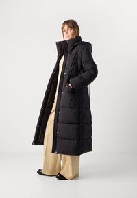 Γυναίκα ντυμένη με μαύρο μακρύ παλτό πούφφερ με ψηλό γιακά, μπεζ παντελόνι σε φαρδιά γραμμή και μαύρα παπούτσια, στέκεται μπροστά σε απλό ανοιχτό φόντο.