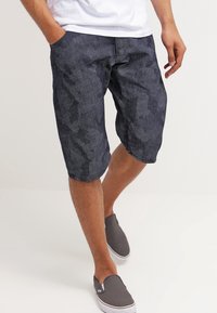 Pantalones cortos oscuros con un corte relajado, que presentan una textura sutil y bolsillos laterales, combinados con unas zapatillas gris claro sin cordones.