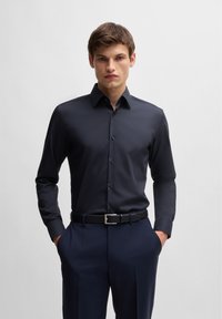 Sötétkék gombos ing sima anyagból, slim fit stílusban, hegyes gallérral és diszkrét fényességgel. Sötét öltönynadrággal és fekete övvel párosítva.