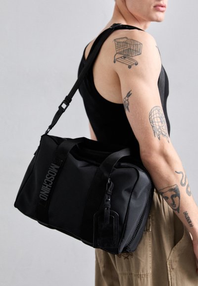 Persoană cu mai multe tatuaje pe brațe poartă o geantă de voiaj neagră Moschino pe umăr, îmbrăcată cu un tricou fără mâneci negru și pantaloni bej.