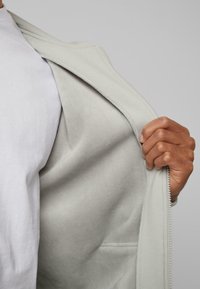 Main tenant une veste gris clair à fermeture éclair ouverte, dévoilant une doublure intérieure douce et lisse ainsi qu'une petite poche intérieure, portée sur une chemise blanche.