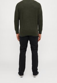 Man med en mörkgrön strukturerad tröja, svarta byxor och svarta sneakers, stående vänd bortåt mot en enkel grå bakgrund.