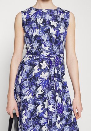 Hihaton tummansininen mekko, jossa valkoisia ja sinisiä kukkakuvioita, istuva vyötärö, ja henkilö, joka pitää mustaa laukkua sivullaan.