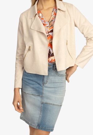 Žena v béžovém krátkém saku se zlatými zipovými kapsami, květované oranžovo-růžové blůze a džínové sukní z patchworku.